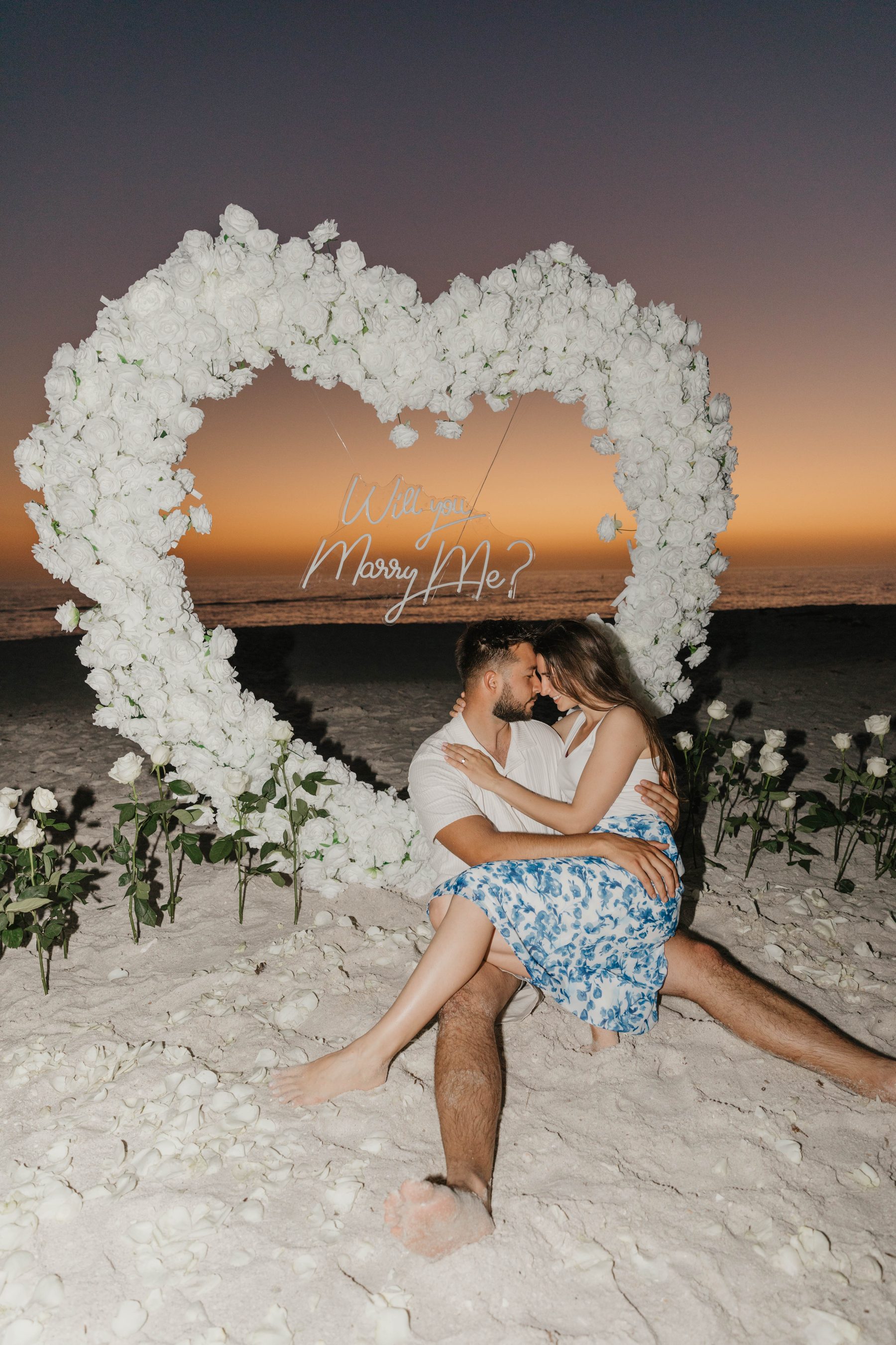 Newly engaged couple embracing inside a white-rose heart arch at dusk — Dejeu Crew engagement photography