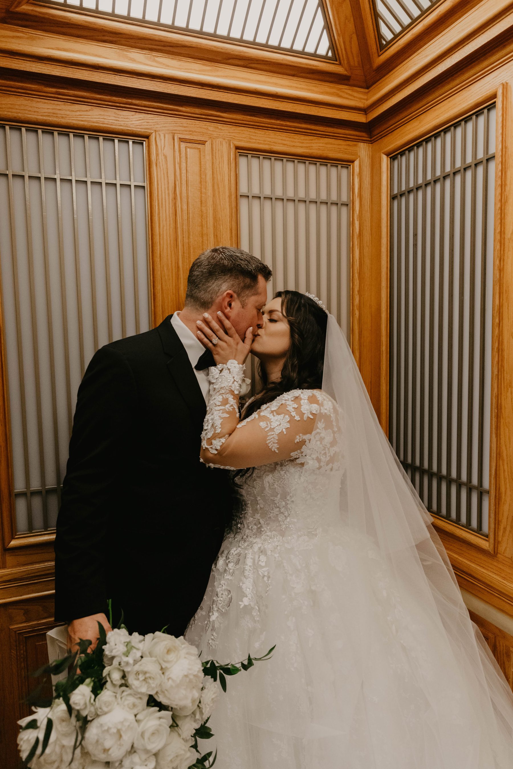 Bride and groom kissing in a warm wood-paneled interior, white rose bouquet in hand — Dejeu Crew wedding photography