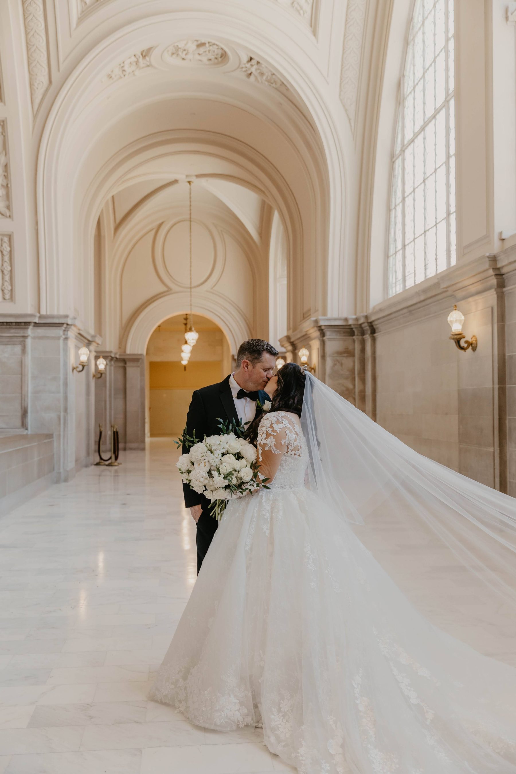 Bride and groom sharing a kiss beneath marble arches, her veil trailing across the floor — Dejeu Crew wedding photography