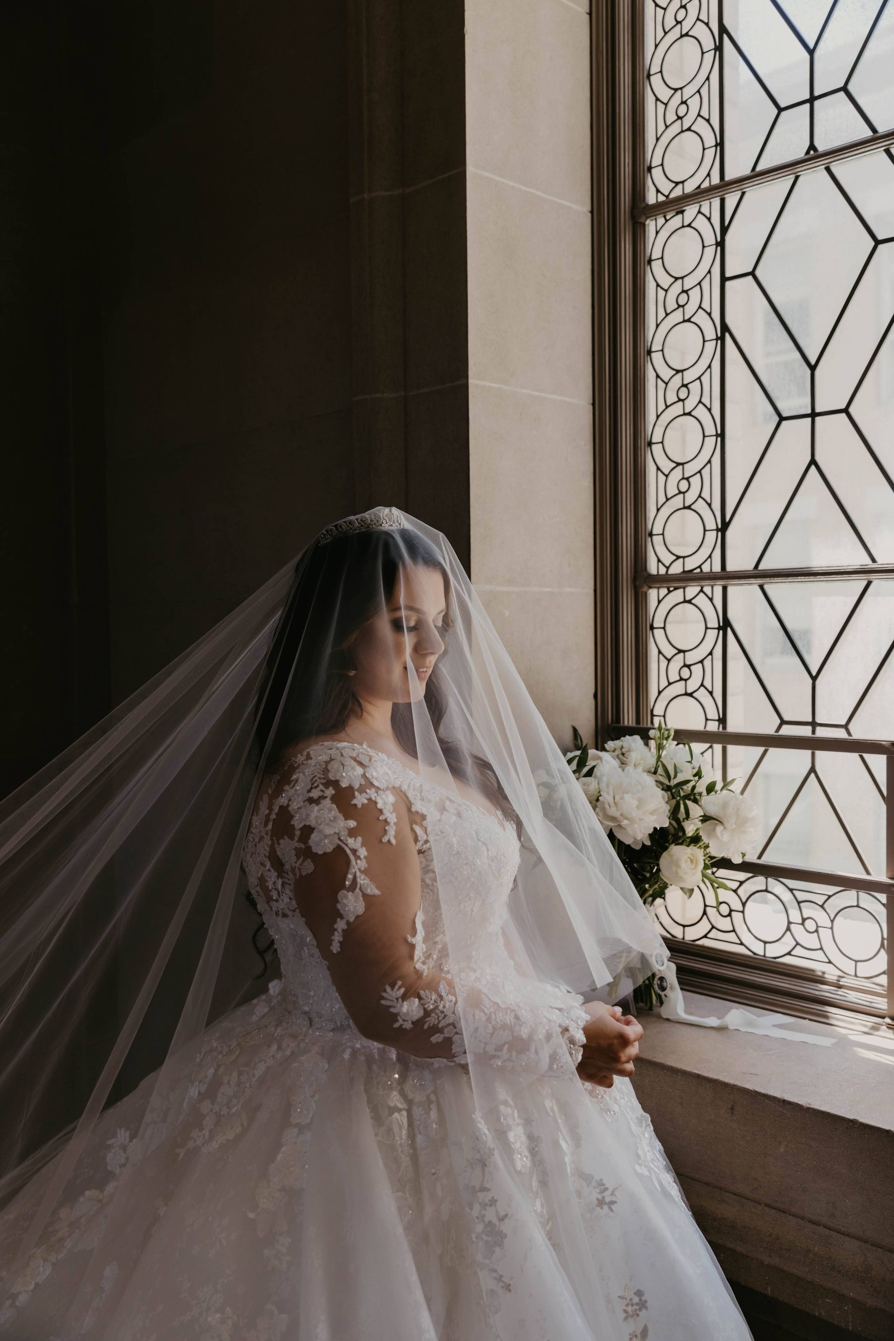 Bride in her veil looking out a tall arched window in soft natural light — Dejeu Crew wedding photography