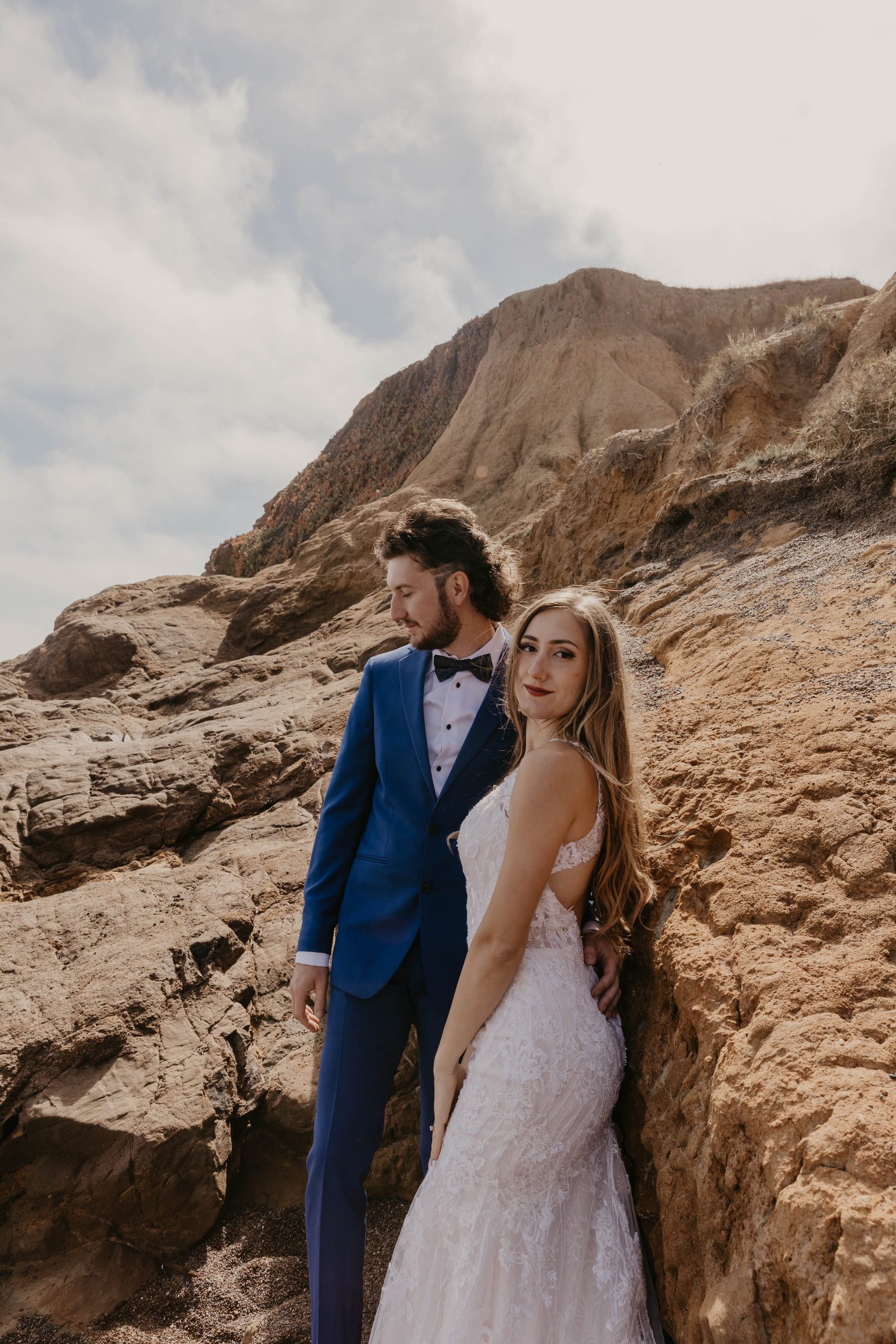 Groom in a blue suit and bride in a lace gown leaning against a sandstone bluff — Dejeu Crew wedding photography
