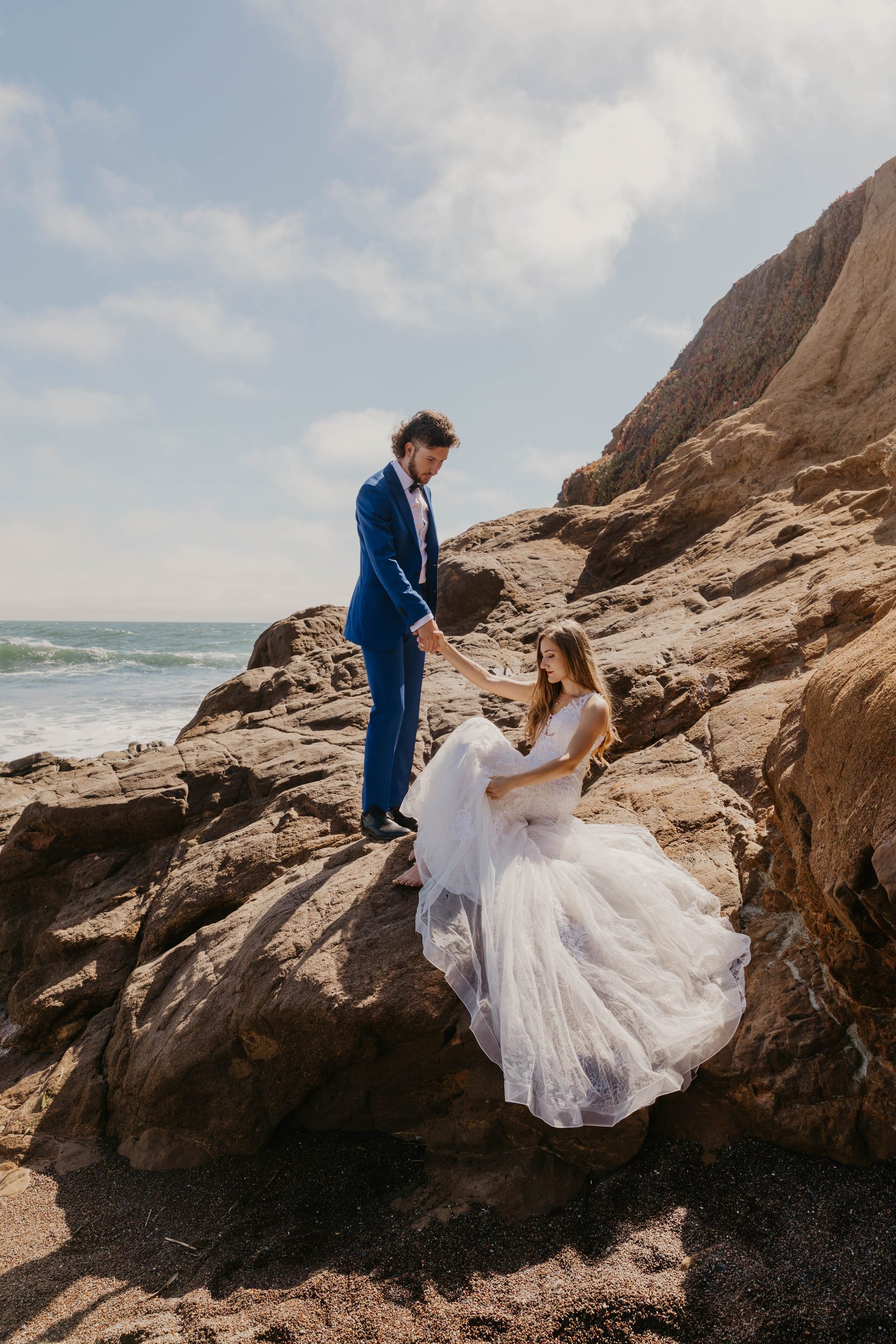 Groom holding the bride’s hand as she sits in her tulle gown on coastal rocks — Dejeu Crew wedding photography