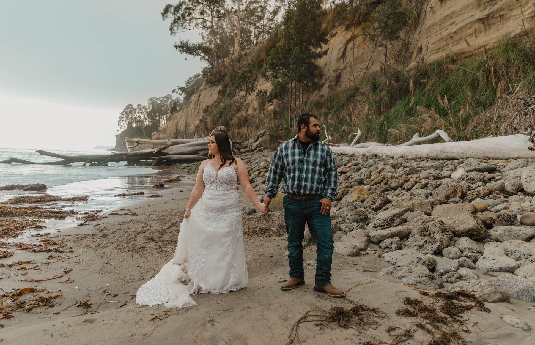 Bride and groom walking hand in hand along a driftwood beach below coastal cliffs — Dejeu Crew wedding photography
