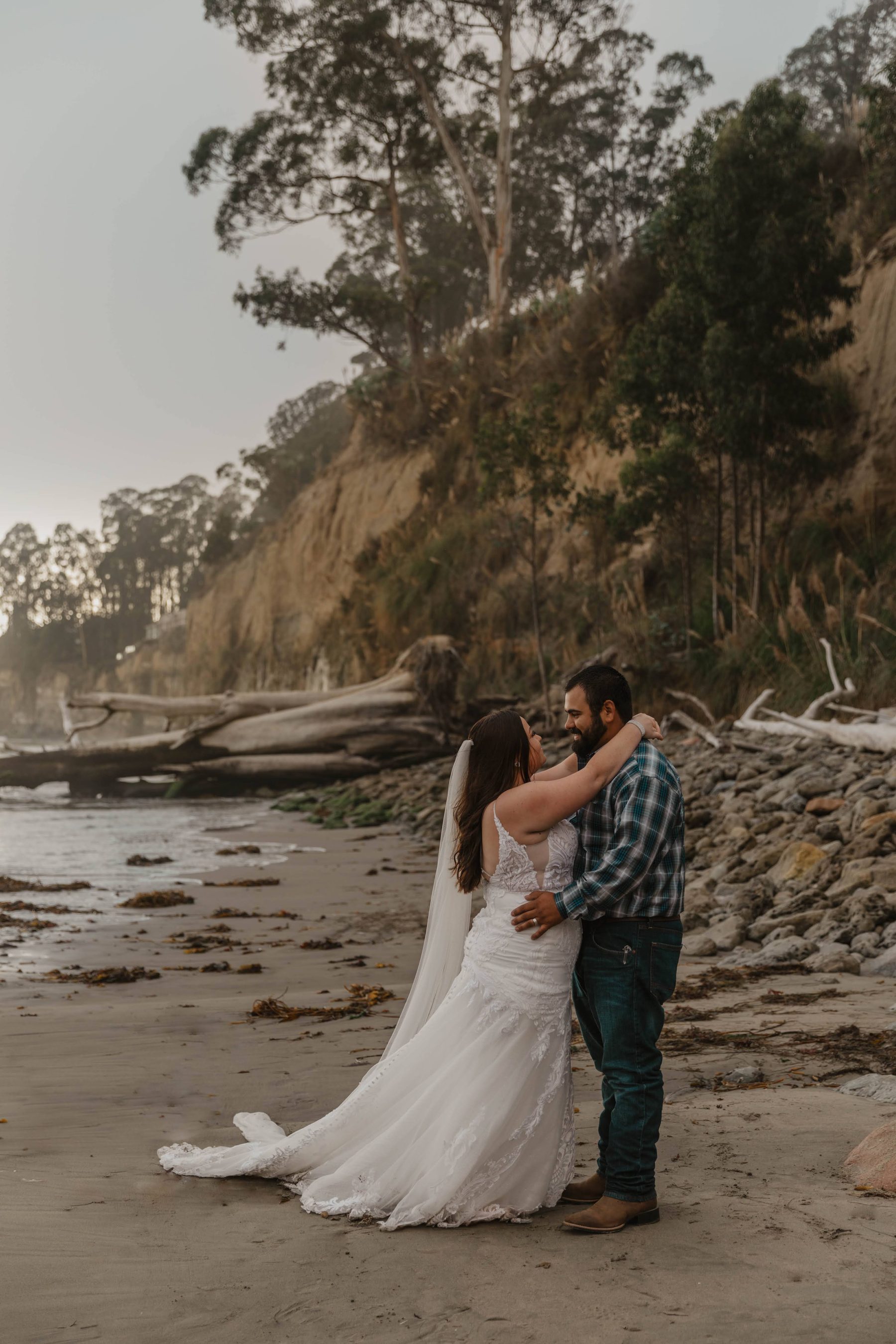 Bride and groom embracing on the beach beneath tree-lined coastal cliffs — Dejeu Crew wedding photography