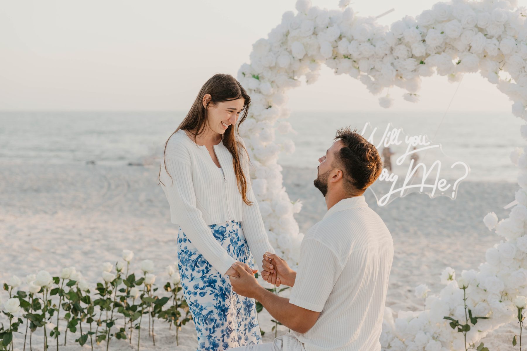 Bride-to-be smiling as her partner proposes in front of a white-rose heart arch on the beach — Dejeu Crew engagement photography