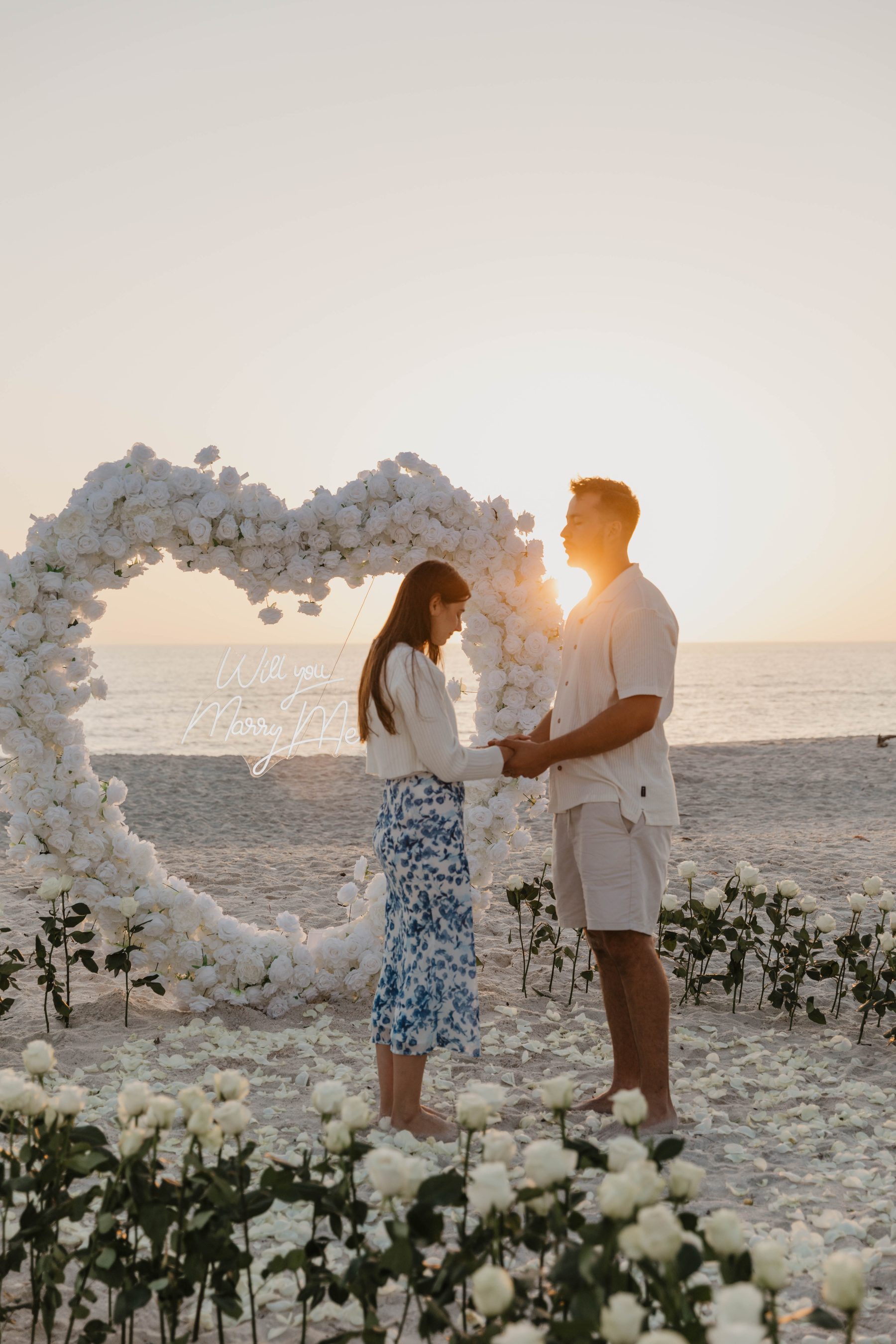 Couple holding hands beside a white-rose heart arch at sunset after a beach proposal — Dejeu Crew engagement photography